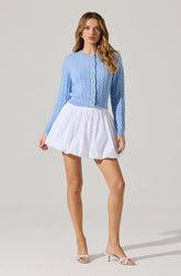 A woman is wearing a light blue, long-sleeved, cable-knit cardigan with pearl buttons down the front. She pairs the cardigan with a short, white, gathered skirt that has a puffy, voluminous shape. She is also wearing open-toed, light-colored high-heeled sandals.